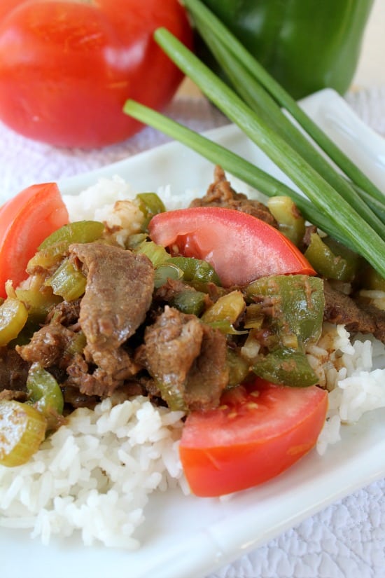 Green Pepper Steak with rice on a white plate with green onions, a bell pepper, and a tomato in the background.