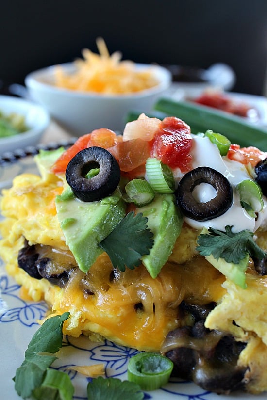 An omelette on a white plate with blue trim that has cheese, olives, avocado, sour cream, green onions, and salsa on it.