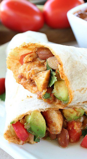 Awesome Chicken Burritos - a burrito cut in half showing the ingredients, sitting on a white plate with two tomatoes in the background.