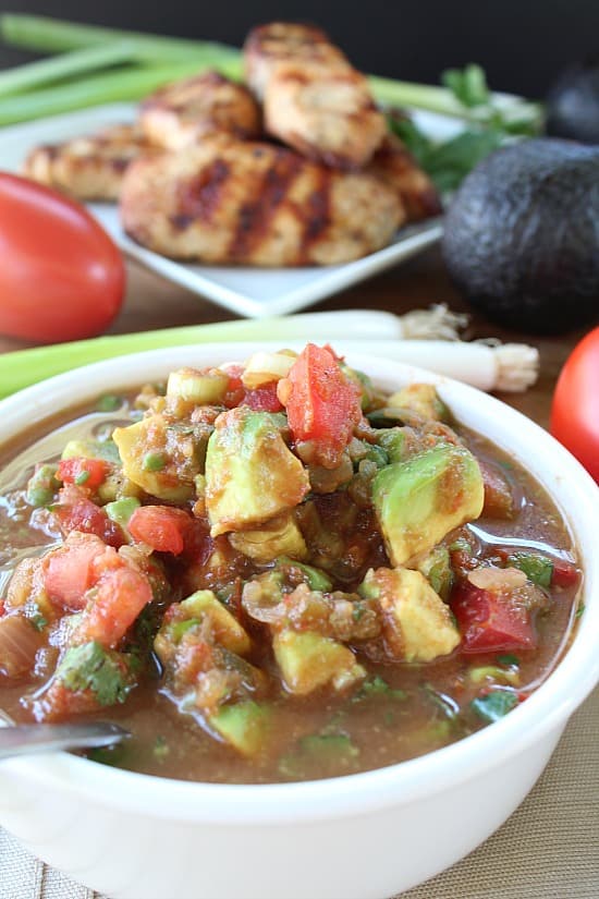 A white bowl full of chunky salsa with chicken strips in the background along with avocado, tomatoes, green onions.