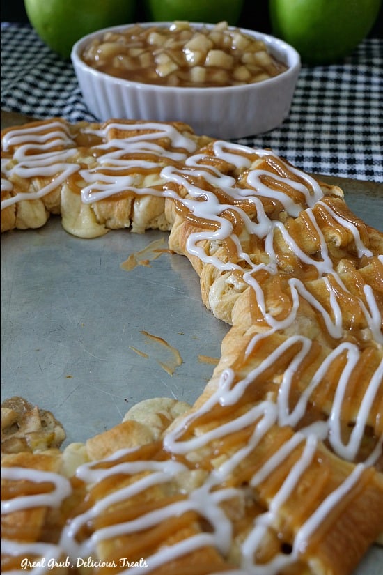 Cream Cheese Caramel Apple Crescent Ring is filled with apples and cheesecake, drizzled with glaze and caramel with a white bowl of apple filling sitting on a black and white checkered towel.