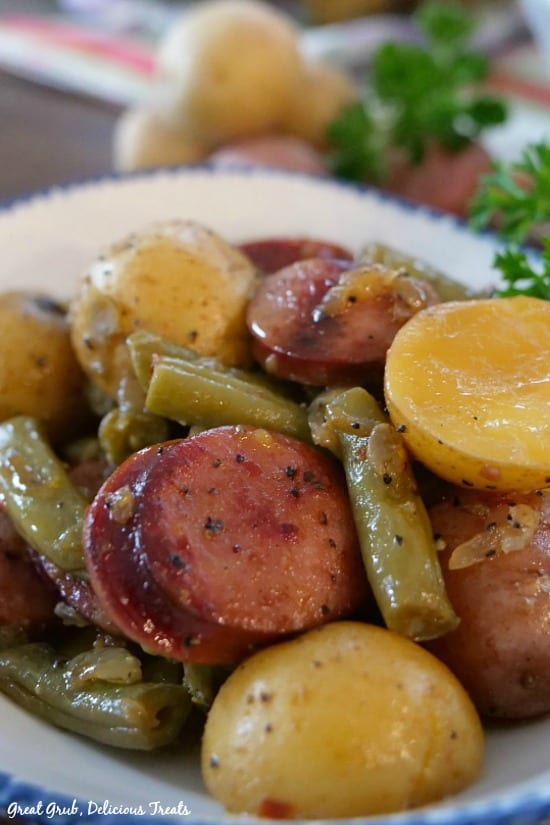 A close up photo of a serving of sausage, green beans and potatoes in a white bowl with blue trim.