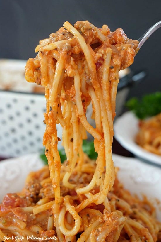 A forkful of Italian Sausage Spaghetti Bake being held up over a serving bowl of spaghetti bake with another bowl and the white and black polka dot baking dish in the background.