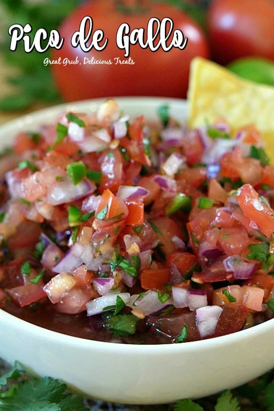 Chopped tomatoes, onions, cilantro, peppers in a white bowl with a tortilla chip in the salsa with two tomatoes in the background.