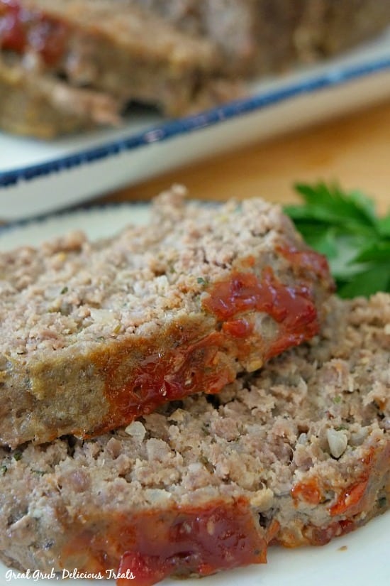 Easy Meatloaf Recipe - two slices of meatloaf on a white plate with more slices of meatloaf in the background on a white plate with blue trim.