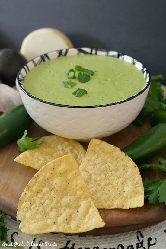 A white bowl with black trim filled with Creamy Jalapeno Cilantro Salsa with tortilla chips in the foreground and jalapenos and cilantro, onions in the background.