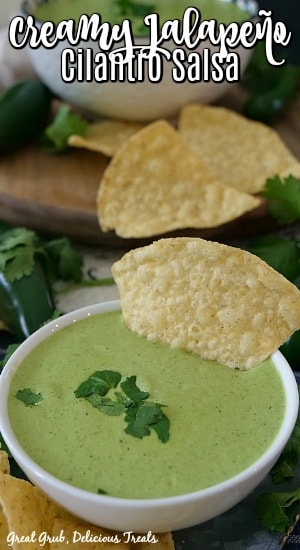 Creamy Jalapeno Cilantro Salsa in a white bowl with a tortilla chip in the salsa with chips, jalapenos and cilantro in the background