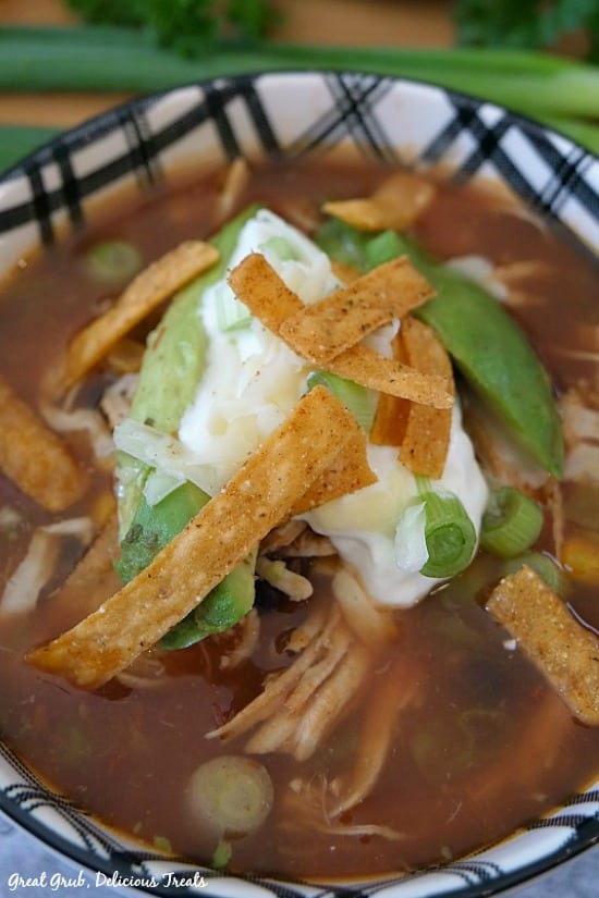 Best Chicken Tortilla Soup in a white and black bowl, garnished with tortilla strips, sour cream, avocado and green onions.