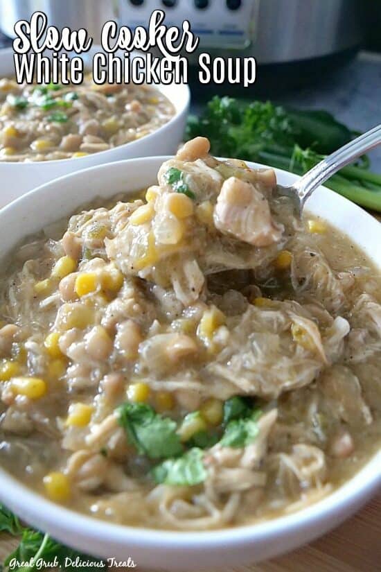 Slow Cooker White Chicken Soup in a white bowl with chicken, corn, beans, and cilantro with another white bowl in the background with the chicken soup and a crockpot for decoration. 