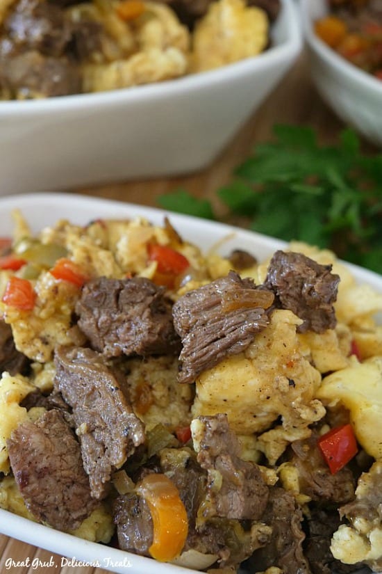 Beef Fajita Breakfast Scramble - a white bowl filled with beef fajita meat, scrambled eggs, peppers and onions, cheese with two more white bowls in the background filled with the breakfast scramble.