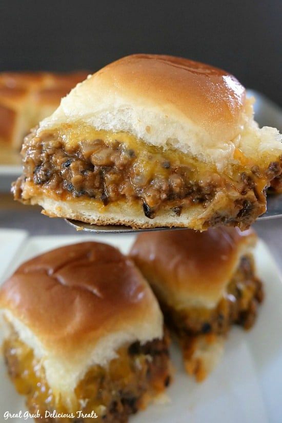 A cheesy beef slider on a spatula being held up in front of the camera for a close up shot with more sliders underneath on a white plate.