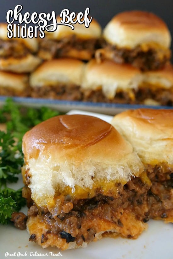 Cheesy beef sliders - a ground beef mixture and cheese between Hawaiian rolls, baked. There are 2 on a white plate with a stack of sliders in the background.
