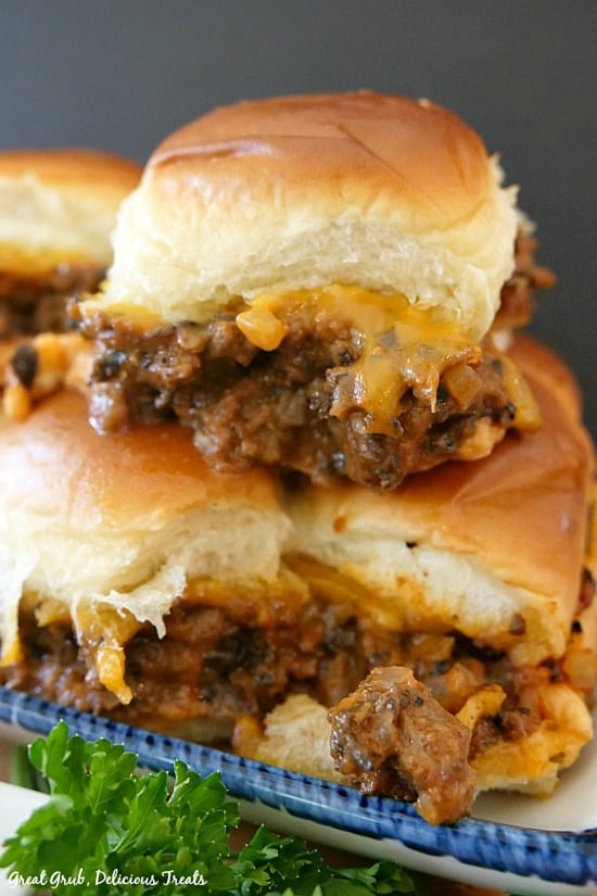 A white plate with blue trim with 4 cheesy beef sliders on it, loaded with ground beef, black olives, cheese and more, baked in between Hawaiian rolls.