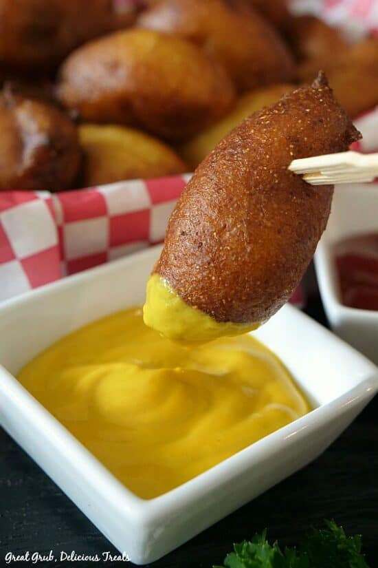 A mini corn dog being dipped into mustard that is in a small white bowl with a basket of mini corn dogs in the background.