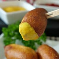 three mini corn dogs with one dipped in mustard and a small white bowl in the background with mustard.