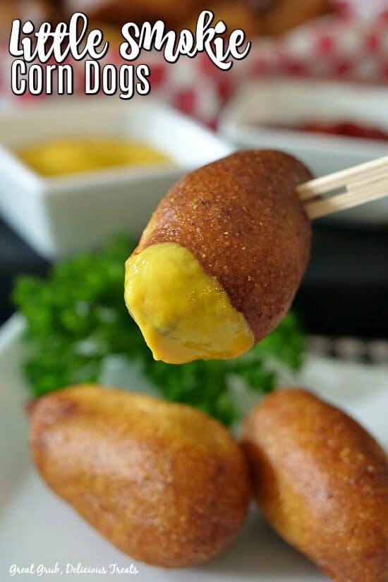 three mini corn dogs with one dipped in mustard and a small white bowl in the background with mustard.