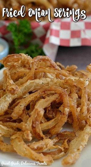 Fried Onion Strings on a white plate with a basket filled with onion rings in the background.