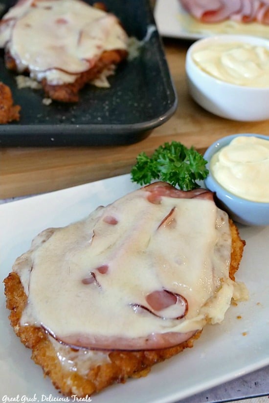 A piece of breaded, fried chicken with melted Swiss cheese and ham on top placed on a white plate with a baking sheet in the background with another piece of chicken, also with two white bowls with dipping sauce inside.