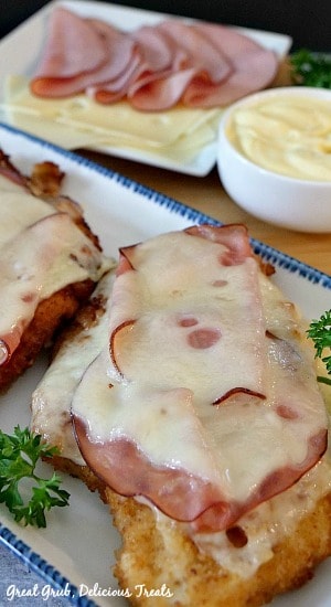 Malibu Chicken is chicken breasts, breaded and fried, then topped with Swiss cheese and ham. This shows two pieces of Malibu chicken on a white plate with sliced ham and cheese in the background on a white plate with a dipping sauce in a white bowl.