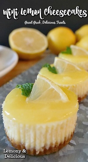 Mini lemon cheesecakes lined up on a silver tray, all with a small lemon wedge and mint on top, lemons in the background.
