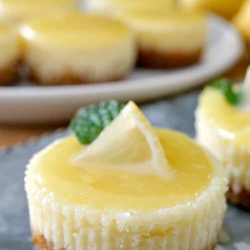 A mini lemon cheesecake with a small lemon wedge and piece of mint on top with mini cheesecakes on a white plate in the background.