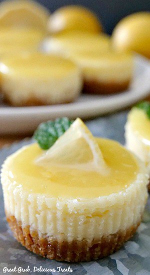 A close up phot of a mini lemon cheesecake with a lemon wedge and a piece of mint on top with other cheesecakes and lemons in the background.