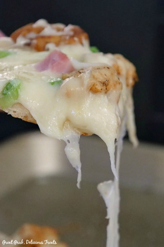Chicken Cordon Bleu Alfredo Pizza - a slice of pizza being held up showing the melted cheese dripping of the sides with chicken, diced ham and green onions on top of the pizza slice.
