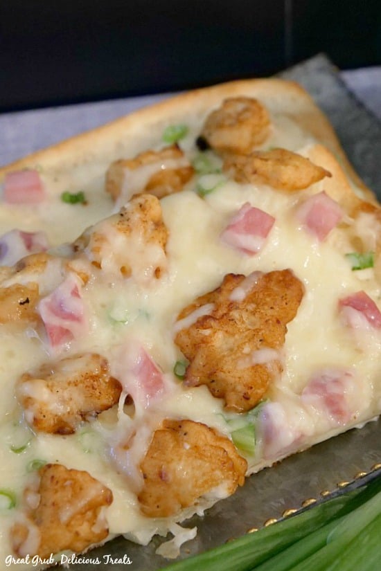 Chicken Cordon Bleu Alfredo Pizza - a piece of pizza with breaded fried chicken, diced ham, green onions, two types of cheese on a silver tray.
