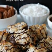Coconut pecan macaroons piled up on a white plate with small white bowls of coconut flakes, pecans, and chocolate in the background.