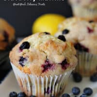 A photo of a blueberry muffin on a white plate with blue trim and a lemon and another muffin in the background.