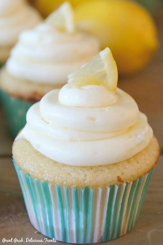 Delicious Lemon Cupcake topped with cream cheese frosting and a small lemon wedge with cupcakes and lemons in the background.