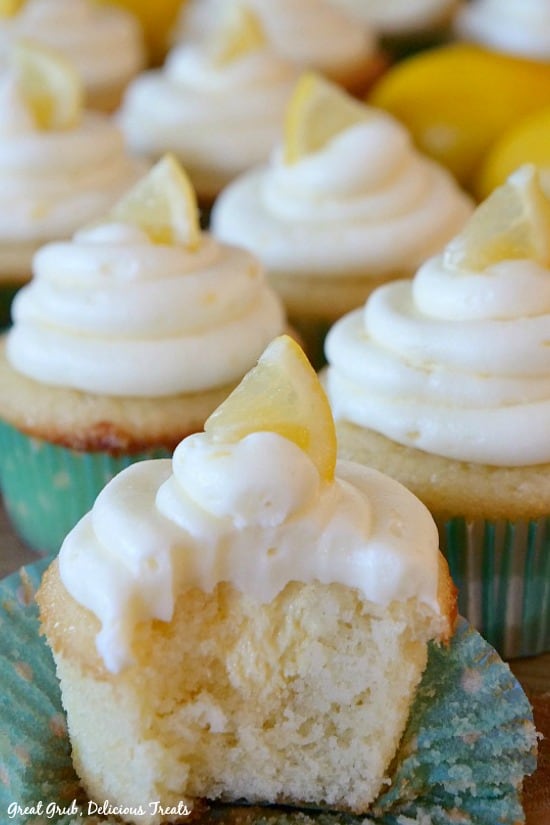 A Delicious Lemon Cupcake with a bite taken out, showing the lemonade cream cheese filling and the sweet lemon cream cheese frosting with cupcakes and lemons in the background.
