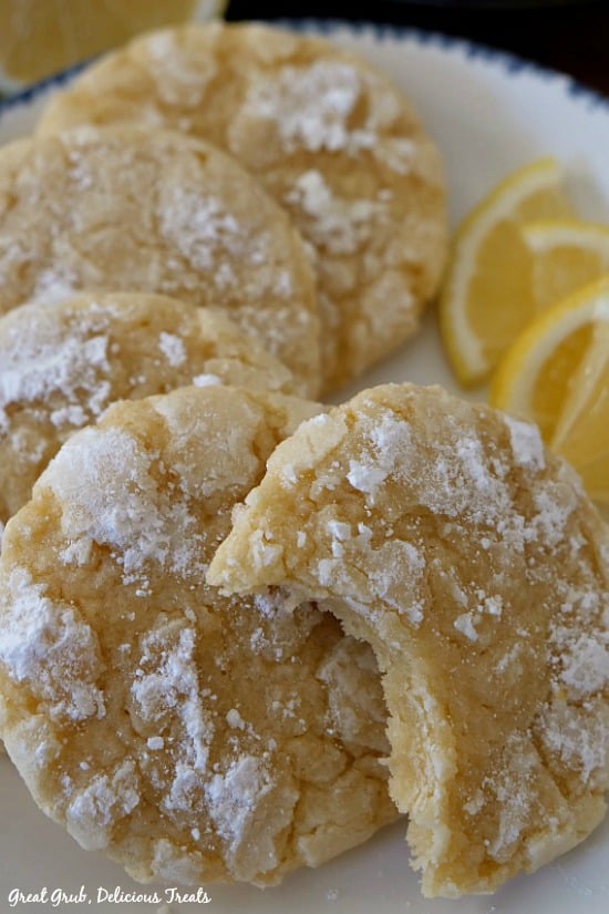A close up photo of 5 lemon crinkle cookies on a white plate with a blue trim with lemon wedges on the side.