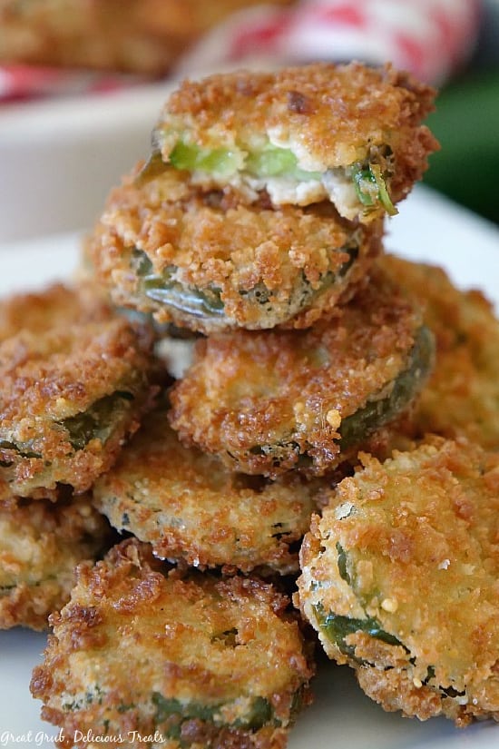  A pile of Deep Fried Crispy Jalapenos stacked up on a white plate with one of the jalapenos with a bite taken out of it showing the sliced jalapeno surrounded by the crunchy coating.