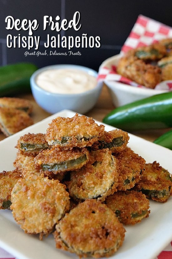 Deep fried jalapeños on a white plate with a small blue bowl on ranch dressing and jalapenos in the background.