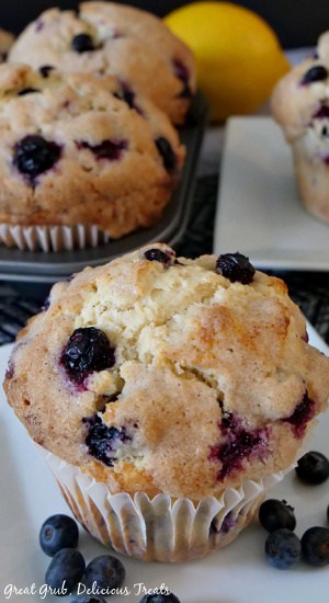 A close up photo of a blueberry muffin on a white plate with another plate in the background and a muffin pan with 2 blueberry muffins in it. 