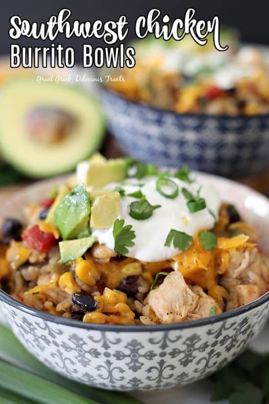 A grey and white patterned bowl filled with rice, black beans, corn, seasoned chicken, tomatoes, avocado, shredded cheddar cheese, sour cream, and cilantro with a avocado half and another burrito bowl in the background.