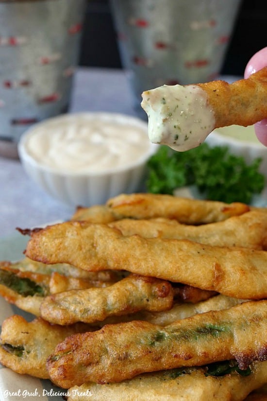 A close of photo of a stack of fried green beans, with one green bean dipped in ranch dressing and a small white bowl of ranch dressing in the background.