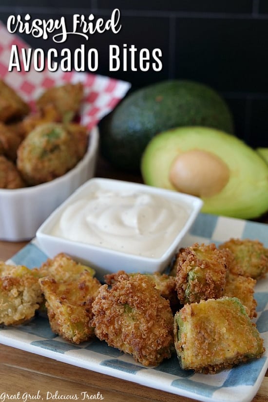 A light blue and white checkered plate with 9 crispy fried avocado bites on it with a white square bowl with ranch dressing in it with an avocado and more avocado bites in the background.
