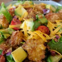 Close up photo of a crispy chicken salad with fried chicken bites, avocado, tomatoes, cheddar cheese, and chunks of bacon.