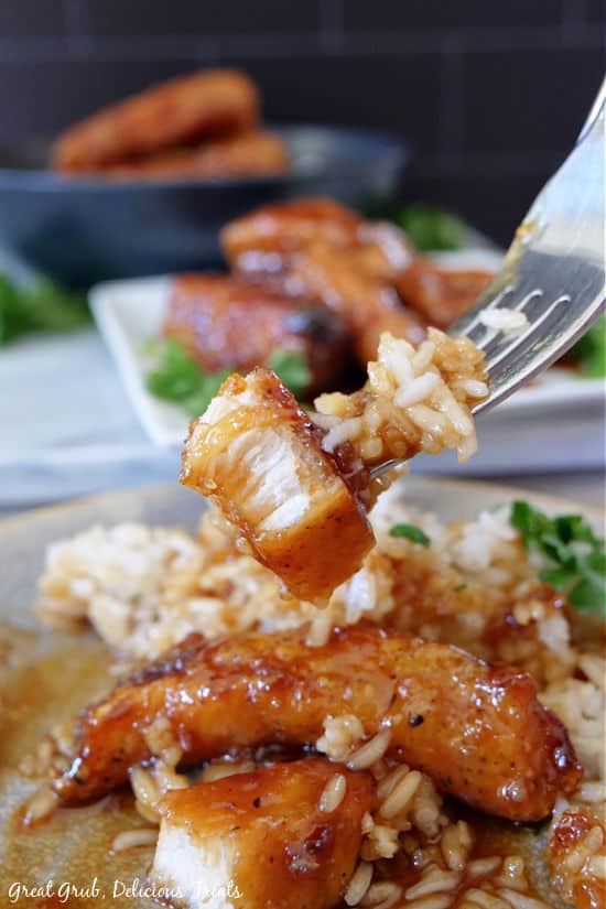 A bite of Lemon Honey Glazed Chicken on a fork held up above a serving of this recipe on a grey plate.