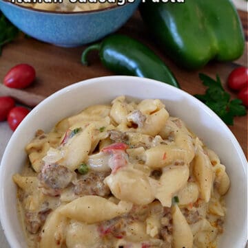 Creamy Italian sausage pasta in a white bowl with a blue bowl of pasta in the background and a bell pepper, a jalapeno, and parsley in the background.
