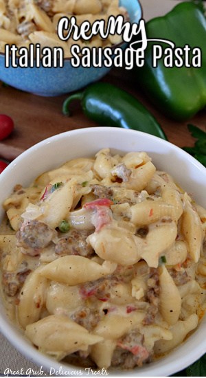 Creamy Italian sausage pasta in a white bowl with a blue bowl of pasta in the background and a bell pepper, a jalapeno, and parsley in the background. 