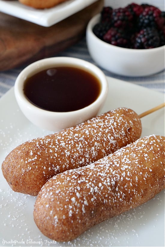 Two pancake sausages on a stick placed on a white plate with a small white bowl filled with maple syrup and another white bowl filled with fresh blackberries.
