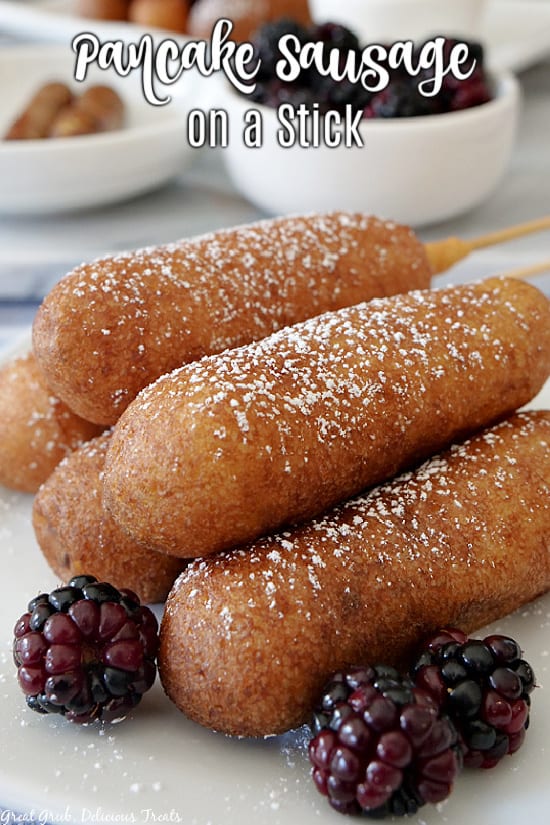 A close up photo of pancake sausage on a stick stacked on top of each other on a white plate with blackberries in the foreground.