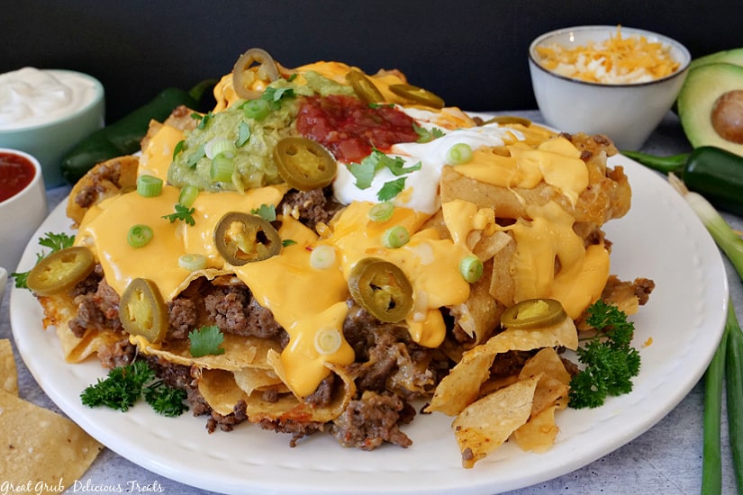 Mile High Nachos stacked up on a white plate with small white bowls holding salsa, sour cream, and shredded cheese, jalapenos, and avocado in the background.l