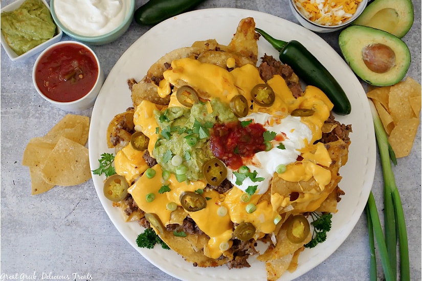 An over view picture of guacamole, sour cream, salsa, shredded cheese in small white bowls, with avocado, jalapenos, green onions and tortilla chips surrounding a white plate loaded with Mile High Nachos.