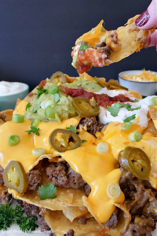 Mile High Nachos stacked on a place showcasing ground beef, nacho cheese, green onions, jalapenos, sour cream, salsa, and refried beans. Two small bowls with sour cream and shredded cheesed are in the background.
