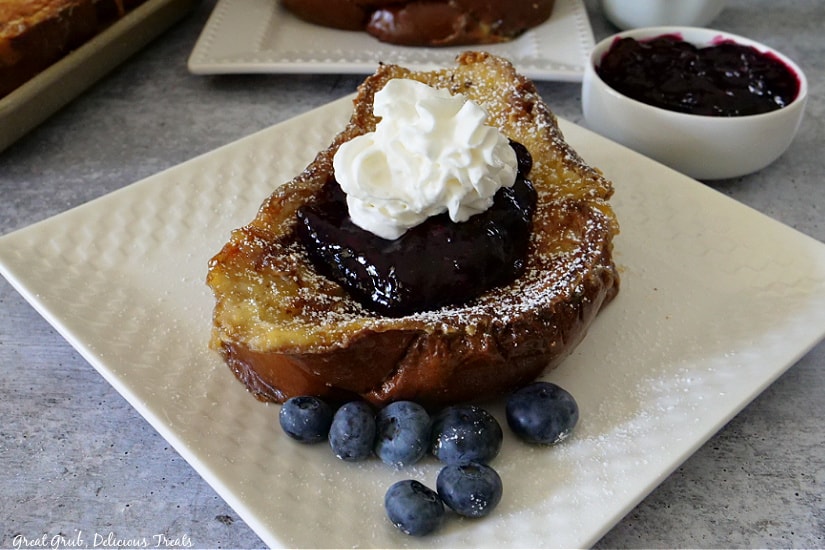 A slice of French toast with blueberry topping placed on a white plate with fresh blueberries on the plate.