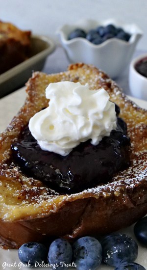 A slice of French toast with blueberry topping on a white plate topped with whipped cream.